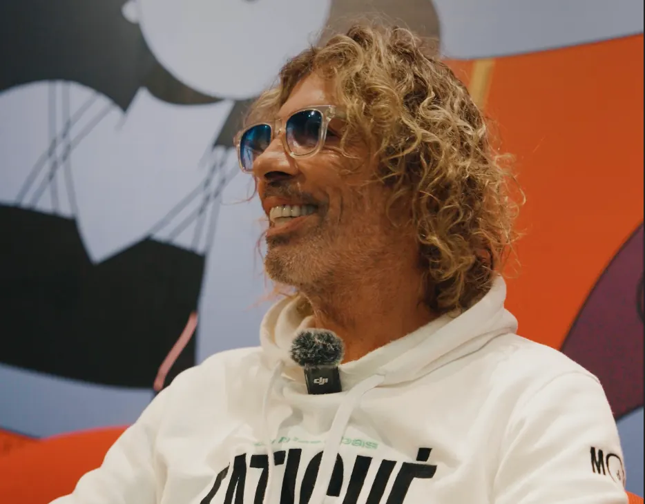 Homme aux cheveux bouclés et lunettes de soleil, interviewé devant une fresque