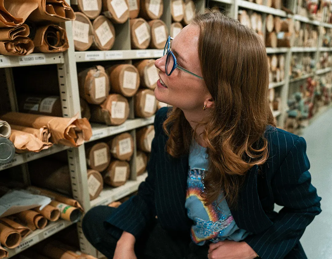 Femme à lunettes en blazer, accroupie entre des rayonnages d’archives