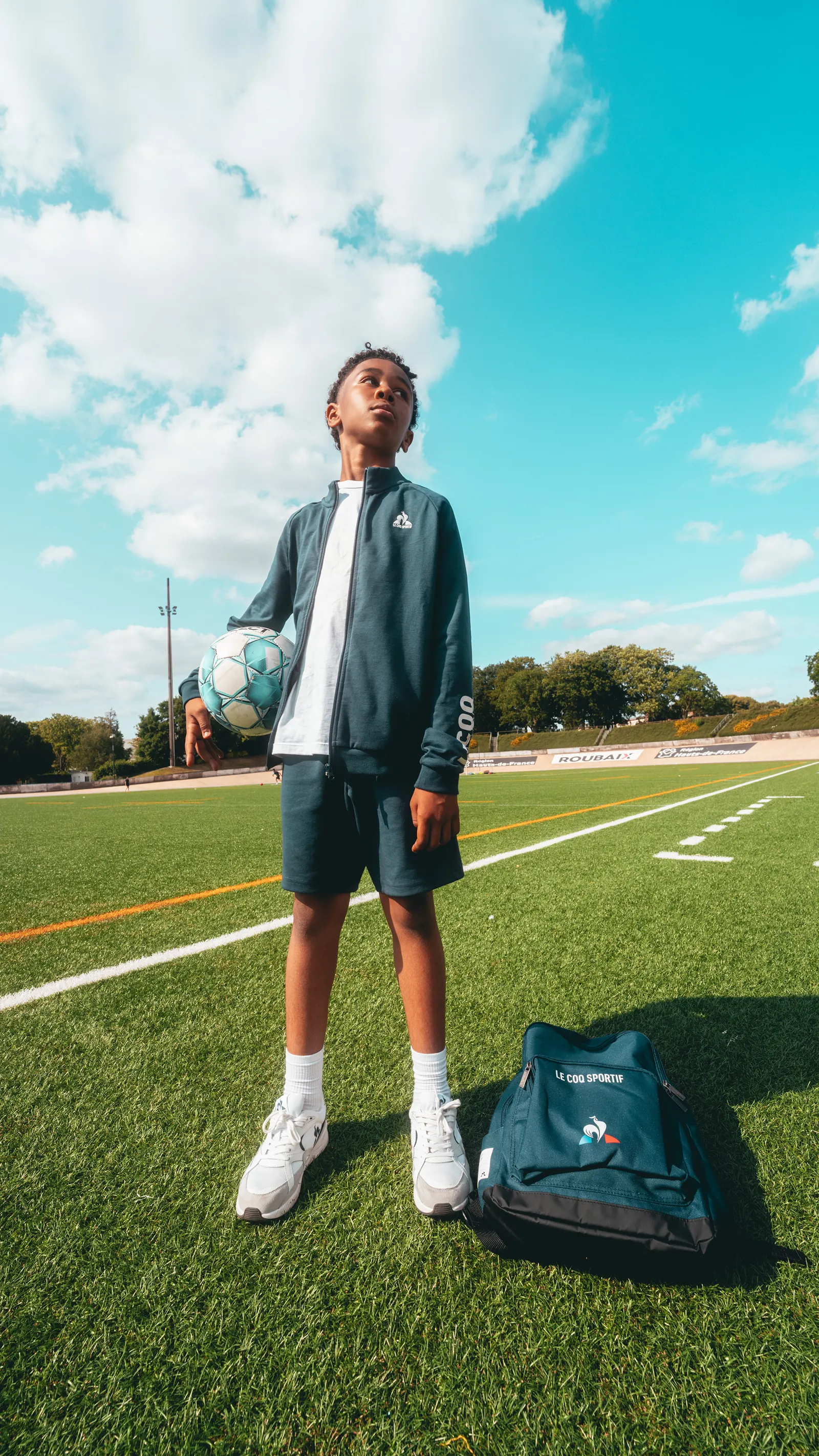 Jeune footballeur en survêtement vert, ballon sous le bras, sur la pelouse